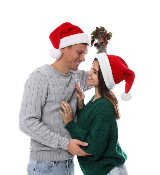 Happy couple in Santa hats standing under mistletoe wreath on white background - Foto, imagen