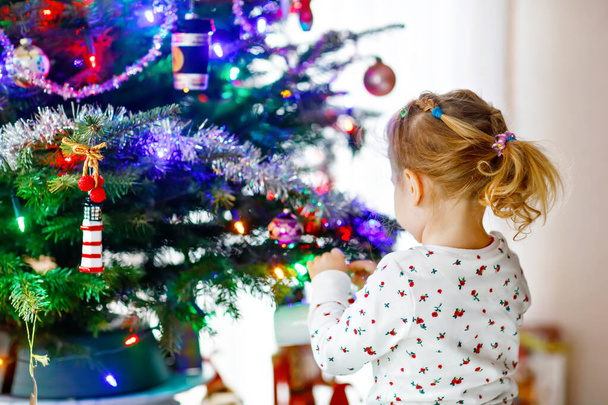 Adorable toddler girl in pajamas decorating Christmas tree with toy in cute hands. Little child in nightwear standing by Xmas tree. celebration of traditional family winter holiday - Photo, Image