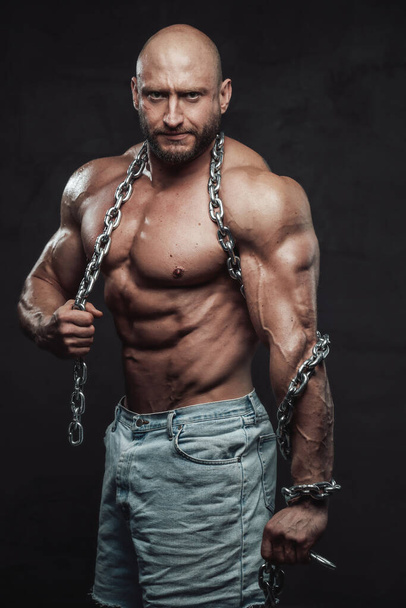 Serious hairless bodybuilder poses with steel chains in dark background - Valokuva, kuva