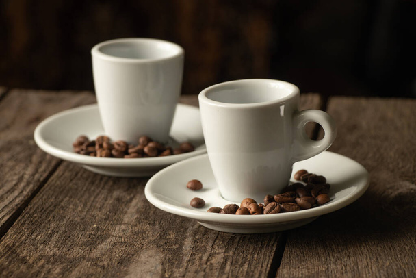 dos tazas blancas y un platillo con granos de café de pie sobre una mesa de madera por la mañana en la cocina - Foto, imagen