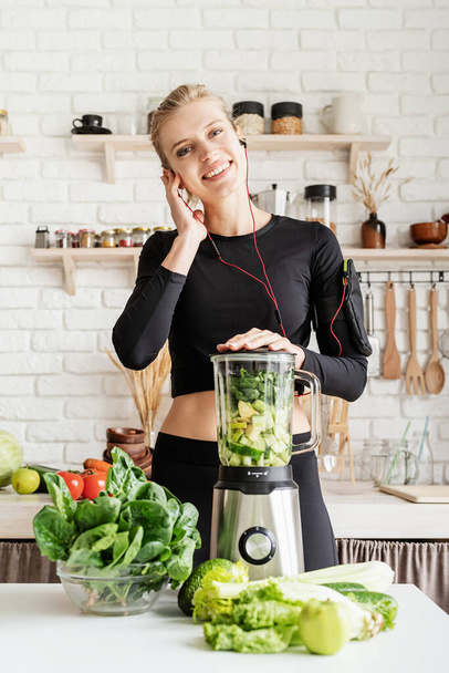 Comida saludable, concepto de dieta. Joven mujer rubia sonriente en ropa deportiva negra escuchando la música y haciendo batido verde en la cocina casera - Foto, imagen