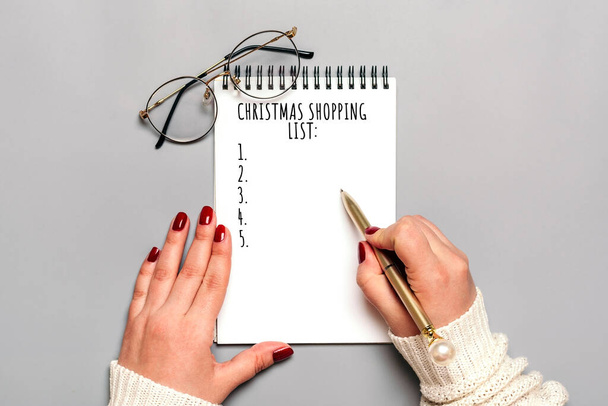 γυναικείο χέρι κρατά στυλό, γράφοντας χριστουγεννιάτικη λίστα αγορών, ιδέες δώρων σε λευκό σημειωματάριο σε γκρι φόντο. - Φωτογραφία, εικόνα