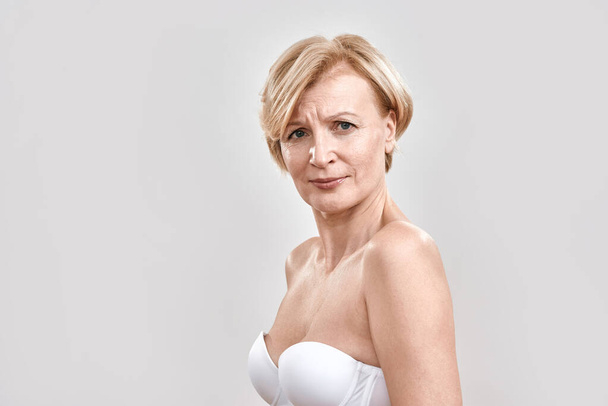 Portrait of beautiful middle aged woman in white bra looking serious at camera, posing isolated against grey background. Beauty concept - Φωτογραφία, εικόνα
