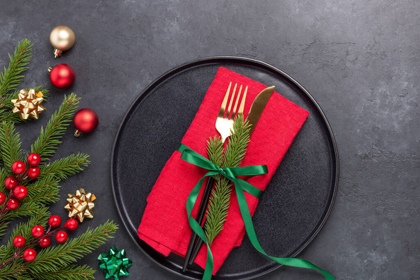 Christmas table setting with black ceramic plate, fir tree branch and gold and red accessories on black stone background. Top view. Copy space - Image - Foto, immagini
