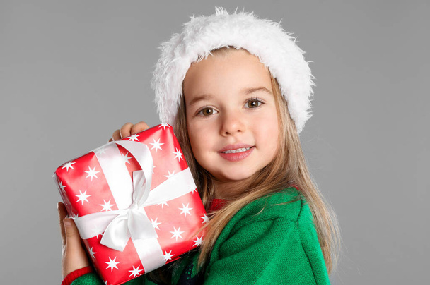 Cute child in Santa hat with Christmas gift on grey background - Фото, изображение