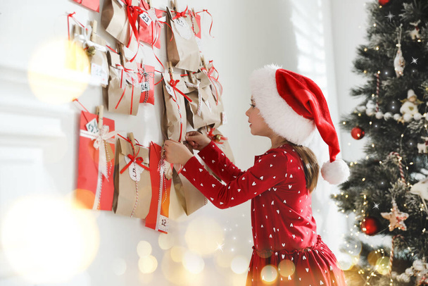 Cute little girl in Santa hat taking gift from Christmas advent calendar at home. Bokeh effect - 写真・画像