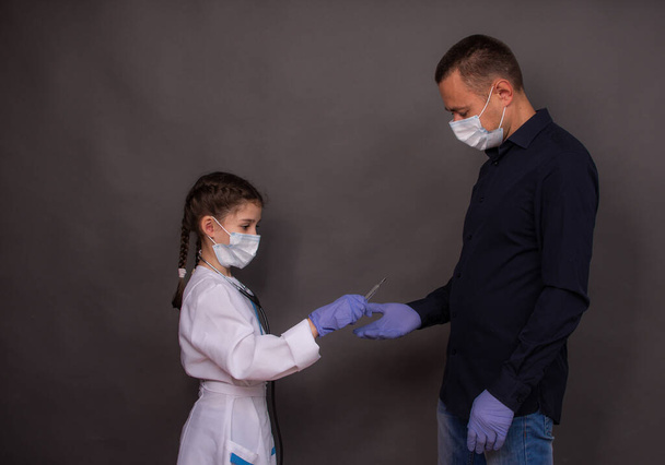 Child girl plays doctor. Adorable kid in medical lab coat gives a medical thermometer to her father. On grey background. Copy space.  - Photo, Image