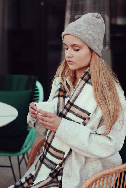  blond long hair beautiful girl in knitted hat and scarf with cup of coffee close up portrait in cafe  - Фото, изображение