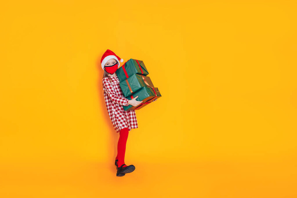 Dziecko trzymające duże pudełka z prezentami. Ma czapkę Mikołaja na głowie i maskę medyczną na twarzy. Boże Narodzenie. Idealne zdjęcie do reklamy świątecznej podczas pandemii. - Zdjęcie, obraz