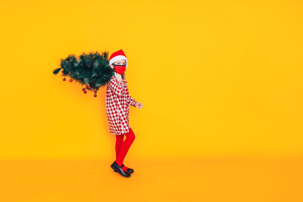 Criança na frente de um fundo amarelo segurando uma árvore de Natal. Humor de Natal. Ela tem um chapéu de Pai Natal na cabeça.. - Foto, Imagem