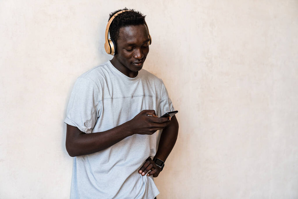 Joven hombre afroamericano con auriculares sosteniendo el teléfono celular mientras está de pie sobre la pared en el interior - Foto, imagen