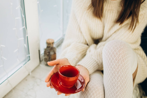 A girl in a white sweater holds a red cup in her hands. Sits on the windowsill against the background of a large window. New year holiday concept - Zdjęcie, obraz