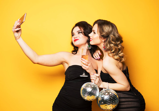 two fashionable young women wearing black cocktail dresses holding disco balls and make a selfie. - Foto, imagen