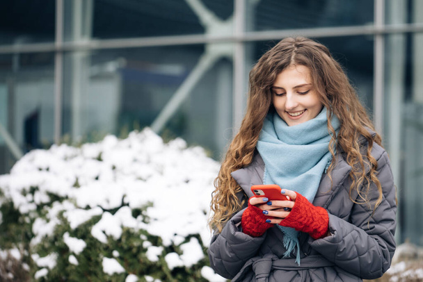 Curly-haired female texting on smartphone standing on street in winter city. Female tapping on cellphone outdoors. Vacation winter outdoor. Happy Young Woman Enjoys Life. - Foto, afbeelding