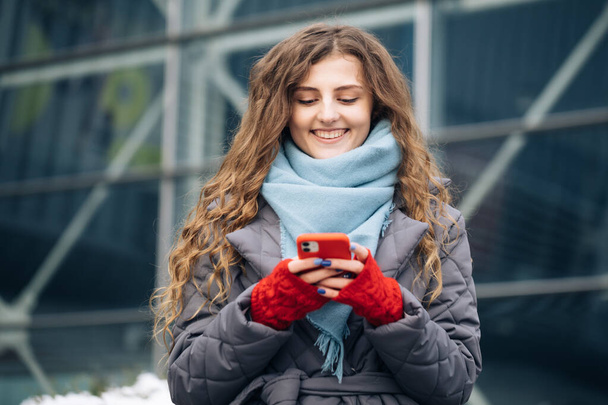 Cheerful Curly-haired Young female texting on smartphone standing on street in winter city on New Year. Female tapping on cellphone outdoors. Online shopping - 写真・画像