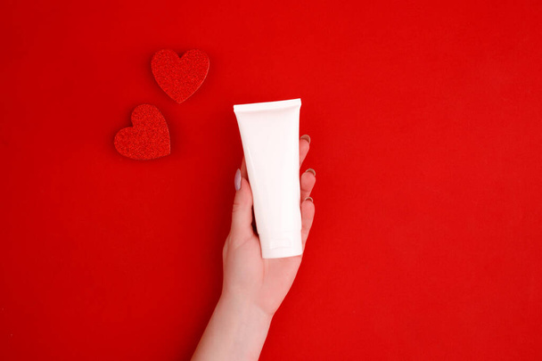 Creme in weiblicher Hand mit Dekor-Herzen auf rotem Hintergrund. Kosmetik. Ansicht von oben. Minimalismus, Attrappe. - Foto, Bild