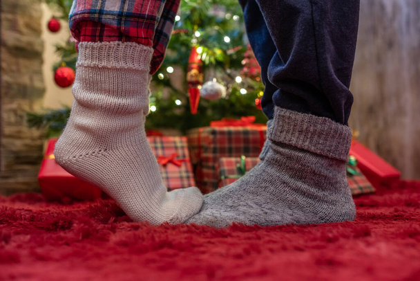 Woman feet standing in tip toe in winter socks on male lags on a fluffy red blanket near a Christmas tree with gifts. Concept  - Fotografie, Obrázek