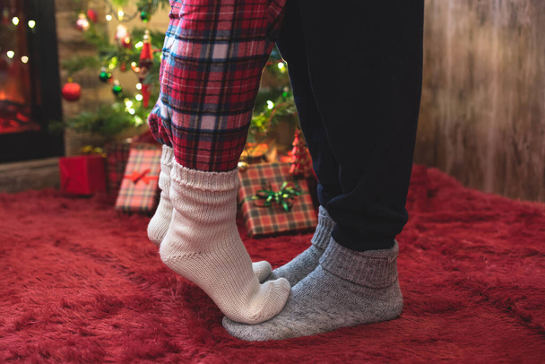 Woman feet standing in tip toe in winter socks on male lags on a fluffy red blanket near a Christmas tree with gifts. Concept  - Zdjęcie, obraz