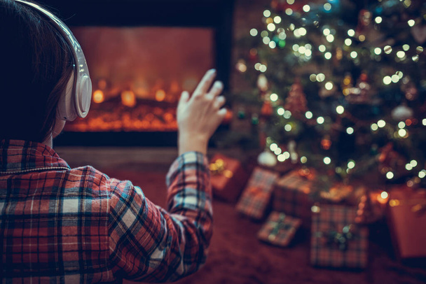 Alone sad woman in headphones sitting and warming at winter evening near fireplace flame and  christmas tree. - Zdjęcie, obraz
