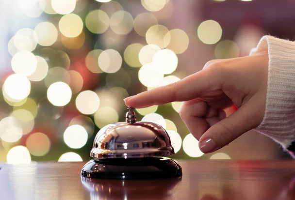 Christmas travel. Hand of guest ringing bell on reception desk of guesthouse and color shining garland on christmas tree on background. Hotel, restaurant. - Fotoğraf, Görsel