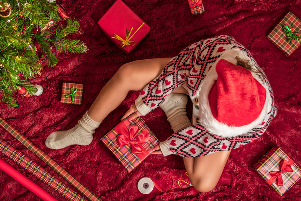Top view of woman in red santa claus hat wrapping christmas gifts boxes for family sitting on fluffy plaid near christmas tree. Flat lay. - Фото, зображення