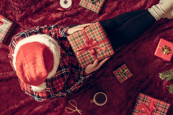 Top view of woman with christmas gift box on knees. Female wrapping gifts for family sitting on fluffy plaid near christmas tree. Flat lay. - Фото, зображення