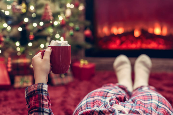 Woman in pajamas with cup of hot cocoa and marshmallow lying and warming at winter evening near fireplace flame and  christmas tree. - Photo, Image