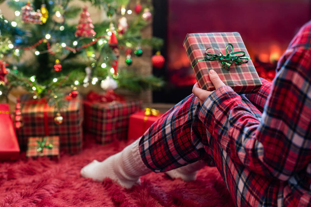 Woman in pajamas with christmas gift box in hand sitting on fluffy plaid near fireplace and christmas tree.  - Zdjęcie, obraz