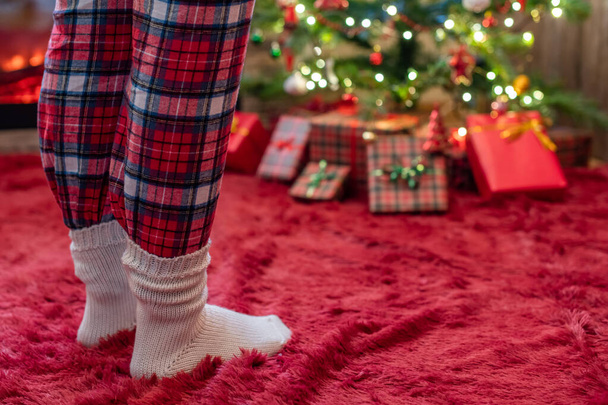 クリスマスツリーの近くに立つパジャマと冬の靴下の女性の足には、午前中に贈り物と暖炉があります。コンセプト - 写真・画像