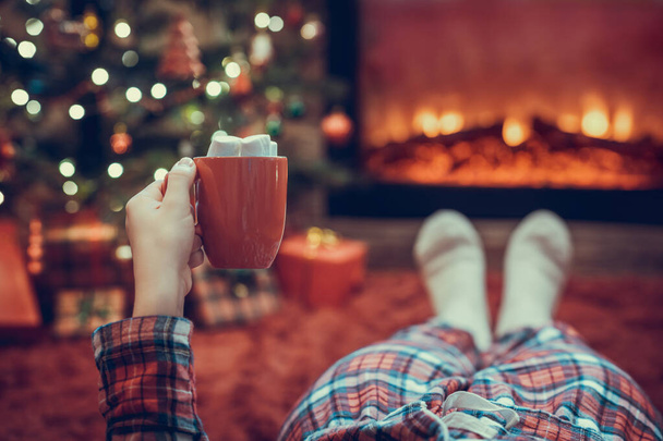 Woman in pajamas with cup of hot cocoa and marshmallow lying and warming at winter evening near fireplace flame and  christmas tree. - Valokuva, kuva