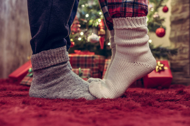 Woman feet standing in tip toe in winter socks on male lags on a fluffy red blanket near a Christmas tree with gifts. Concept  - Фото, зображення