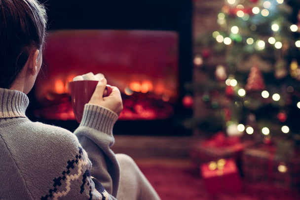 Woman with cup of hot cocoa and marshmallow sitting and warming at winter evening near fireplace flame and  christmas tree. - Zdjęcie, obraz