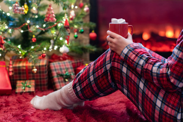 Woman in pajamas with cup of hot cocoa and marshmallow sitting and warming at winter evening near fireplace flame and  christmas tree. - Fotoğraf, Görsel