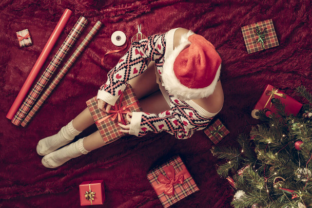 Top view of woman with christmas gift box on knees. Female wrapping gifts for family sitting on fluffy plaid near christmas tree. Flat lay. - Фото, зображення