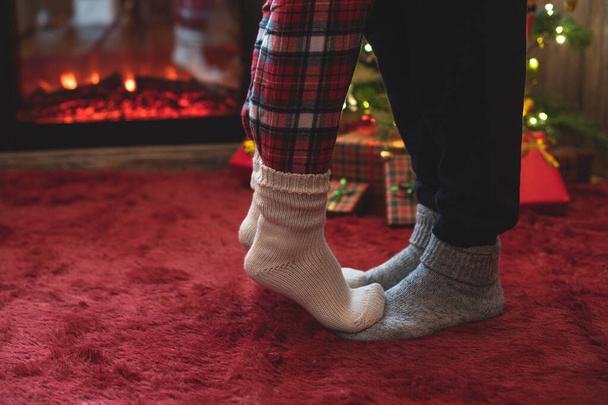 Woman feet standing in tip toe in winter socks on male lags on a fluffy red blanket near a Christmas tree with gifts. Concept  - Фото, зображення