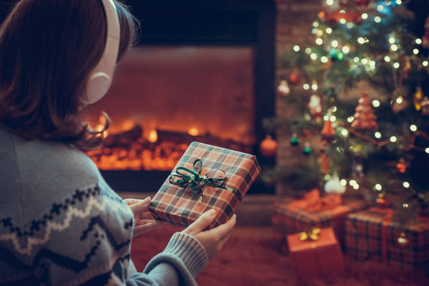 Woman in headphones with christmas gift box in hand sitting on fluffy plaid near fireplace and christmas tree.  - Valokuva, kuva