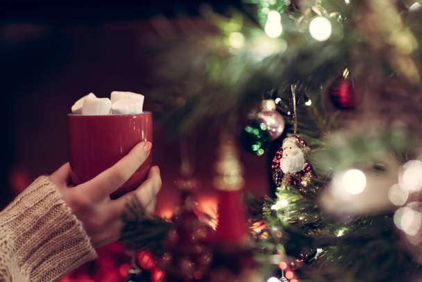 Vrouw hand met kopje warme chocolademelk en marshmallow zit en warmt 's winters bij open haard vlam. Uitzicht door de takken van een kerstboom - Foto, afbeelding