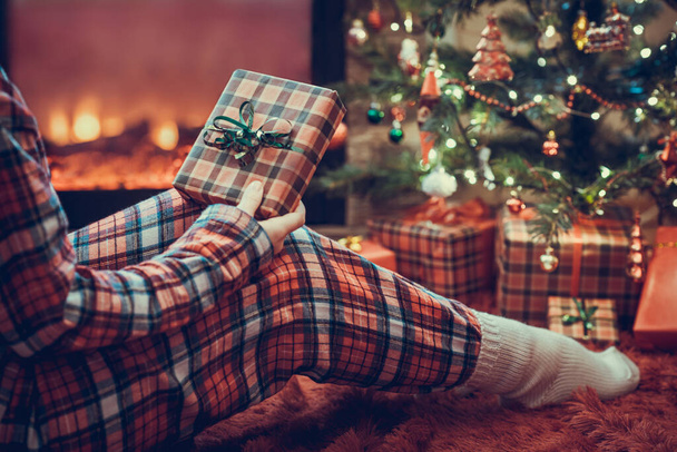 Woman in pajamas with christmas gift box in hand sitting on fluffy plaid near fireplace and christmas tree.  - Foto, Imagen