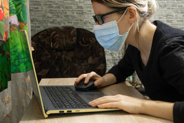 een meisje met een medisch masker zit aan een tafel en draagt een laptop thuis in quarantaine in Oekraïne - Foto, afbeelding