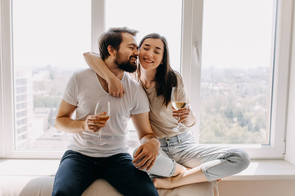 красивая молодая пара с бокалами вина сидит на подоконнике и проводит время вместе дома - Фото, изображение