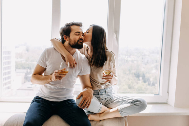 beautiful young couple with glasses of wine sitting on windowsill and spending time together at home - Photo, Image