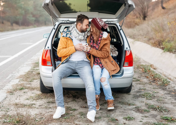 beautiful young couple sitting in car trunk while spending time together outdoors - Photo, Image