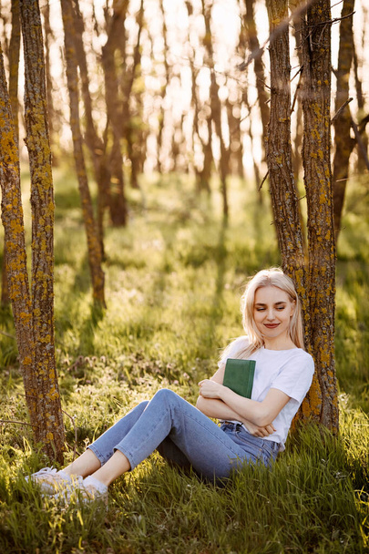 portrait of beautiful young blonde woman reading book while sitting near tree on nature - Photo, Image