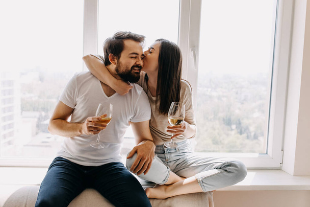 gyönyörű fiatal pár egy pohár borral ül az ablakpárkányon, és együtt töltik az időt otthon - Fotó, kép