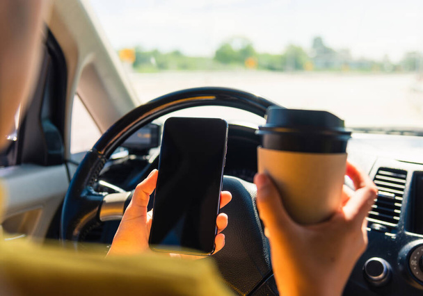 Asian woman drinking hot coffee takeaway cup inside a car and using smartphone blank screen while driving the car in the morning during going to work on highway, Transportation and vehicle concept - Foto, Imagen