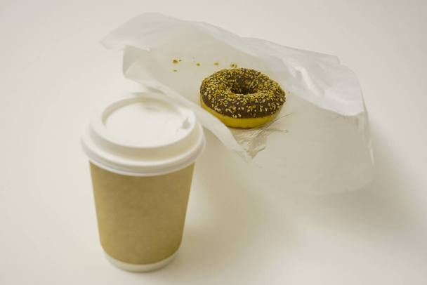 Close Up of one chocolate donut in a paper takeaway bag and a cup of coffee on white table. The delicious deserts covered in powdered nut, bought from a street fast food. - Foto, imagen
