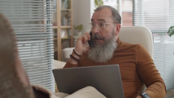 Starší šedovlasý bělošský obchodník sedící na židli s nohama na stole, držící notebook, mluvící po telefonu, poslouchající dobré zprávy, smějící se, chválící mluvčího, ukazující ano gesto rukou - Záběry, video