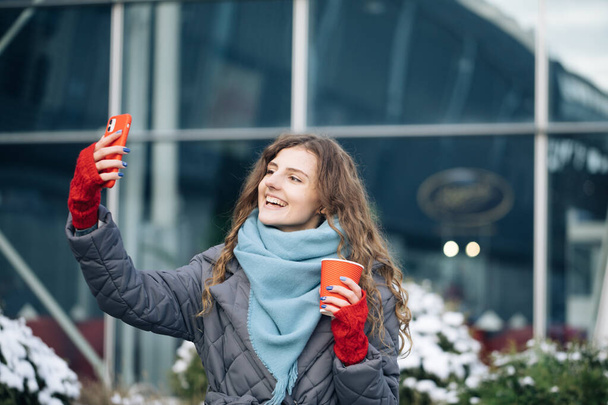 Portrait of happy girl taking selfie holding camera at city street. Modern technology, lifestyle and joyful people concept - Photo, Image