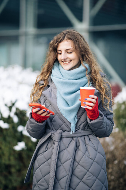 Female portrait. Walking look around on the street city. Being online, social networks. Modern stylish curly woman with cup coffee, uses her smartphone - Foto, immagini