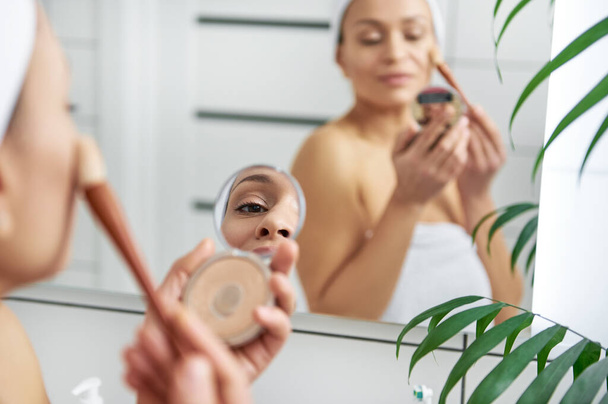 Tükröződés egy fiatal nő tükrében, aki egy kiemelőt rak az arccsontjára. - Fotó, kép